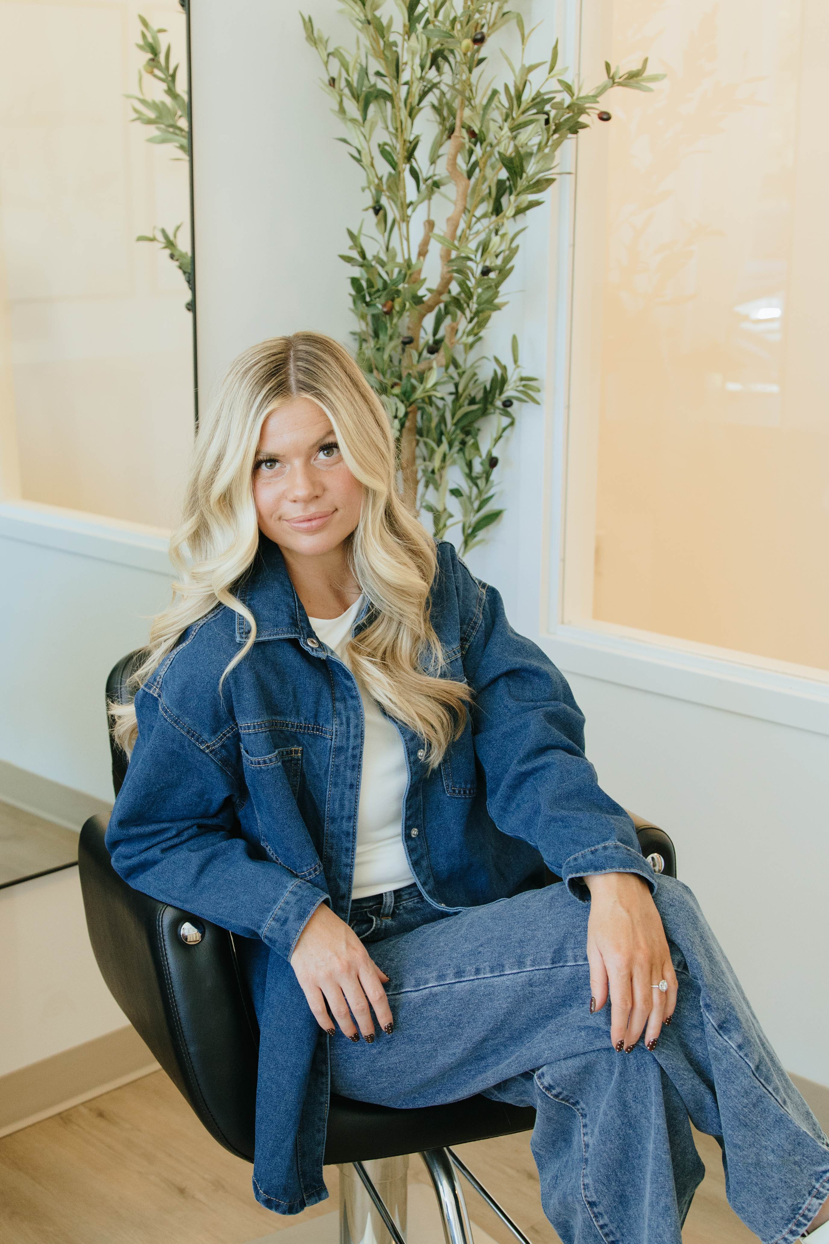 Founder seated in a denim jacket with greenery behind her.
