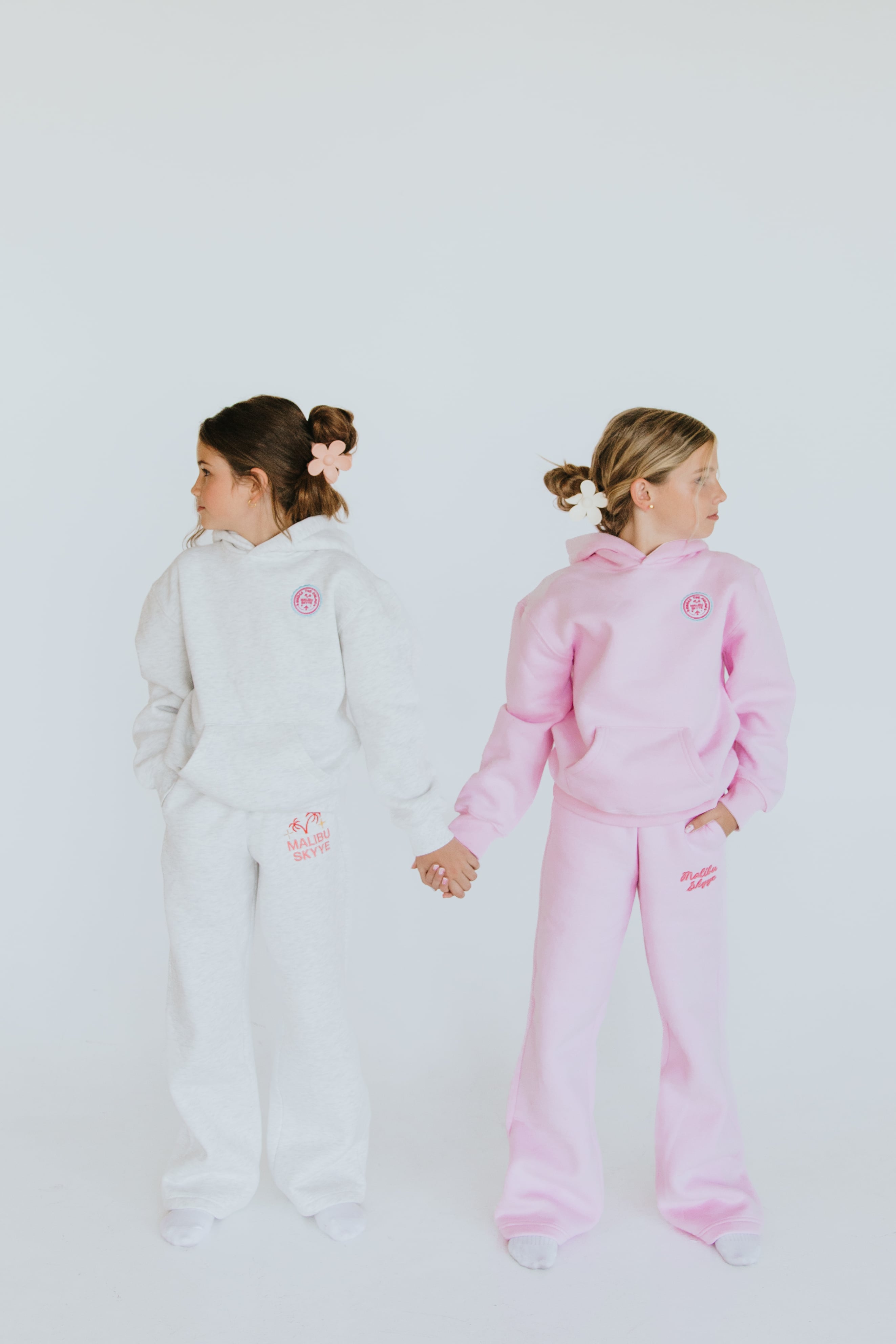 Two models in gray and pink Malibu Skyye sweatsuits holding hands in a bright studio.